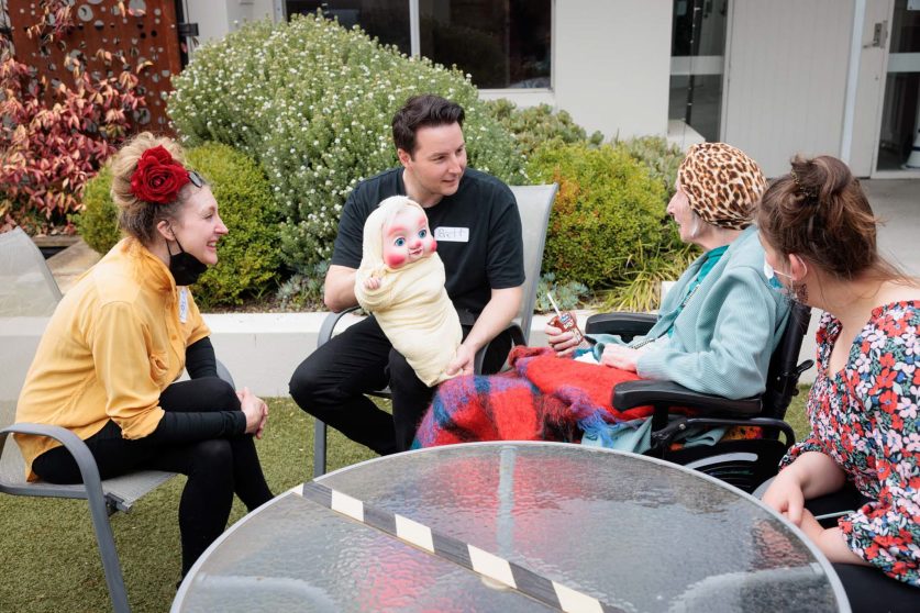 Performers, one holding a puppet, interacts with a resident outdoors