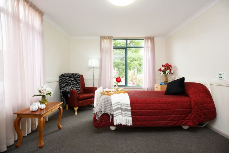 Well-lit room with carpet, desk, single bed, arm chair and flowers.